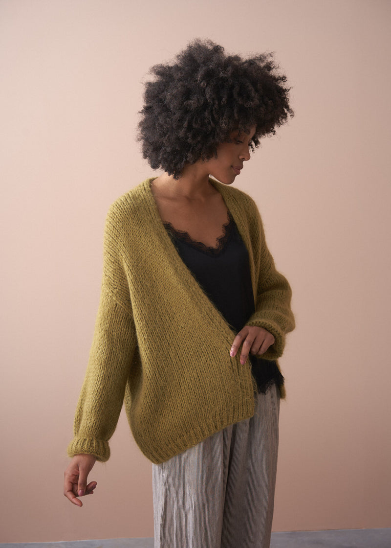 Woman wearing a chunky knit olive cardigan, black satin cami and striped trousers on neutral background