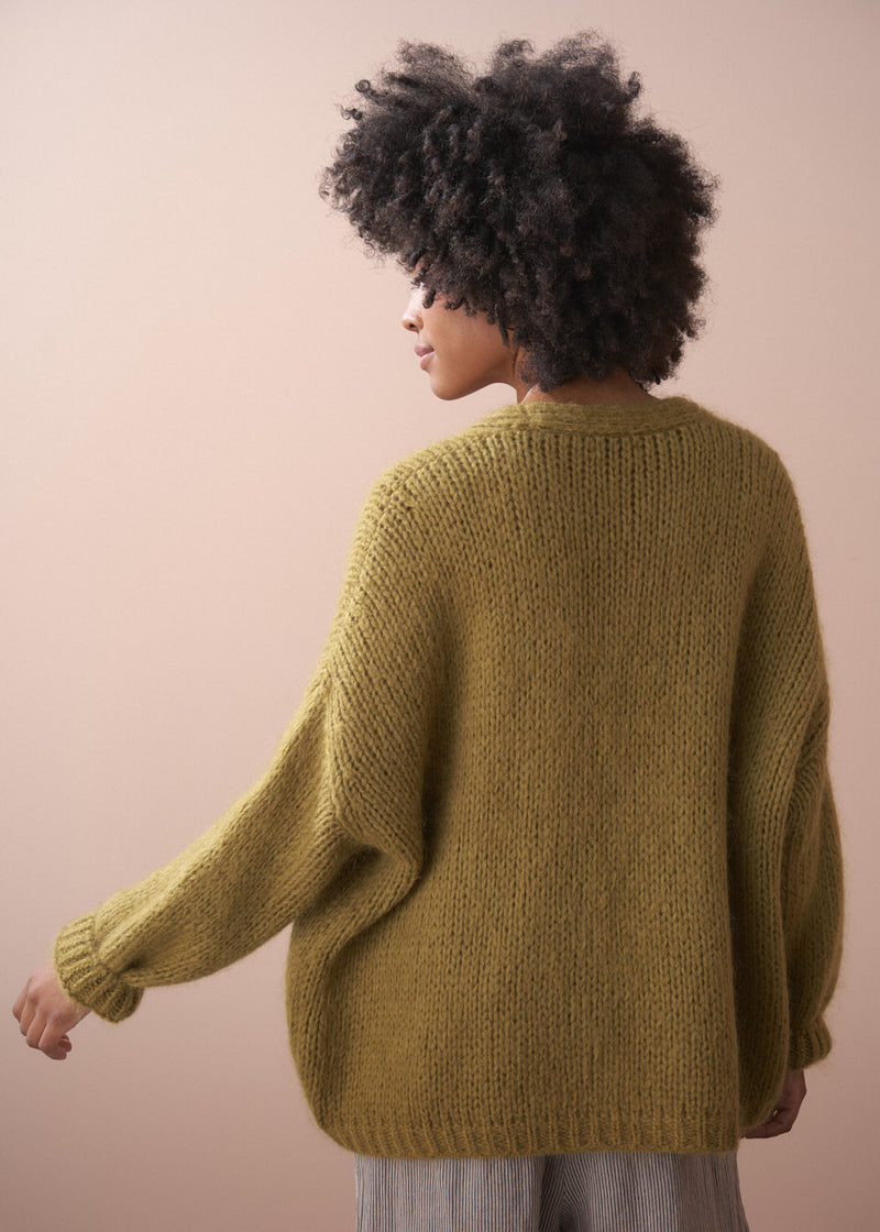 Woman wearing a chunky knit olive green chunky cardigan, facing a neutral background