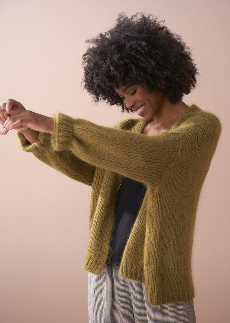 Woman wearing an olive green chunky cardigan, with black satin top and striped trousers.
