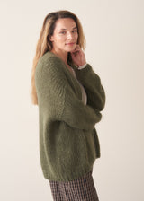 A woman wearing an oversized chunky knit sweater in khaki green with dark checked barrel leg trousers