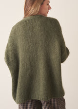 A woman wearing an oversized chunky knit sweater in khaki green with dark checked barrel leg trousers