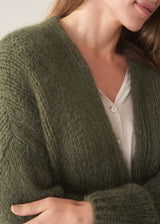 A woman wearing an oversized chunky knit sweater in khaki green with dark checked barrel leg trousers