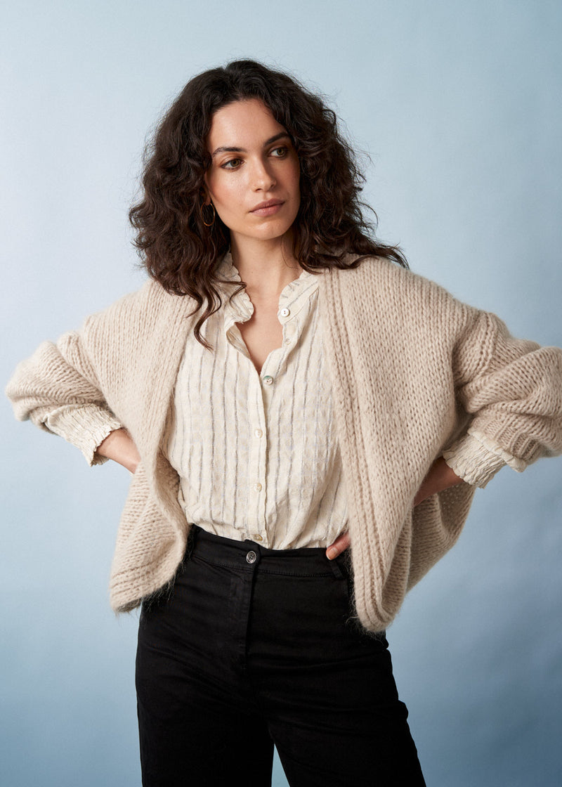 A woman in a soft oatmeal chunky knit cardigan with black trousers, on a neutral background