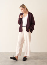 A woman modeling a dark burgundy knit cardigan over a white V-neck top, paired with cream wide-leg trousers and black slip-on shoes, set against a minimalist beige and gray studio background.