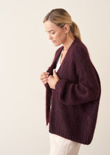 A woman wearing a chunky, fuzzy dark plum purple cardigan over a white V-neck top and white trousers, posing with her arms crossed against a plain off-white wall.