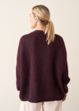A woman wearing a chunky, fuzzy dark plum purple cardigan over a white V-neck top and white trousers, posing with her arms crossed against a plain off-white wall.