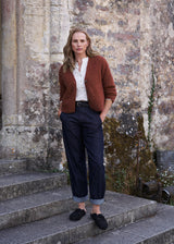 A woman stands on stone steps in front of a weathered stone wall, wearing a rust-brown cardigan over a white top, dark blue jeans, and black slip-on shoes.
