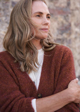 A woman with shoulder-length blonde hair wearing a textured rust-brown cardigan over a white top, posed against a blurred stone wall background.