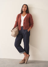 A woman wearing a rust-colored knit cardigan over a white V-neck top and dark blue jeans, holding a beige tote bag against a plain off-white wall.