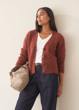 A woman wearing a rust-colored knit cardigan over a white V-neck top and dark blue jeans, holding a beige tote bag against a plain off-white wall.