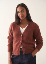 A woman wearing a rust-colored knit cardigan over a white V-neck top and dark blue jeans, holding a beige tote bag against a plain off-white wall.