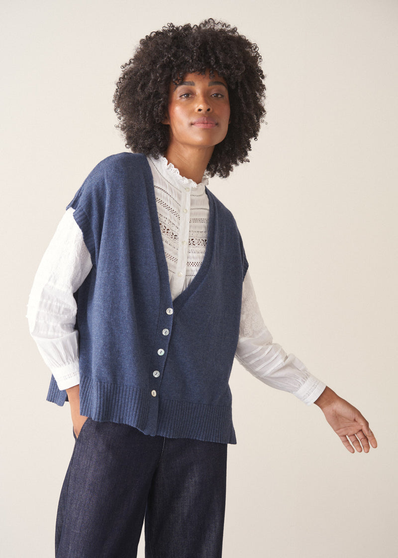 Woman wearing a blue knitted waistcoat, white embroidered top and denim jeans. 