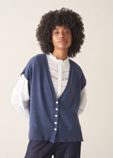 Woman wearing a blue knitted waistcoat, white embroidered top and denim jeans. 