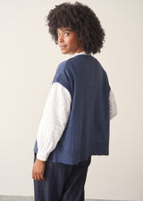 Woman wearing a blue knitted waistcoat, white embroidered top and denim jeans. 