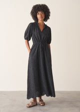 Woman wearing a black maxi broderie dress with deep v-neck and sandals, against a beige background