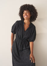 Woman wearing a black maxi broderie dress with deep v-neck, against a beige background