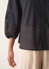 A woman wearing a sheer black embroidered blouse with light linen trousers.
