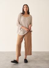 Woman wearing a beige lightweight knitted sweater with camel linen trousers and black clogs against a beige background