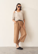 Woman leant against a beige wall, wearing a lightweight beige knitted sweater with camel trousers and black clogs