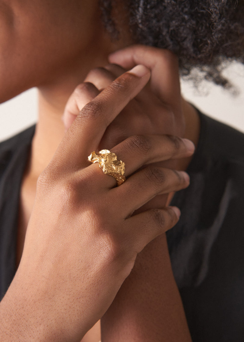 Close-up of a woman's hands clasped together, showing a large, textured molten gold ring on her finger
