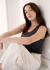 Woman wearing a dark grey jersey tank top with white jeans, leaning against a neutral background