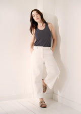 Woman wearing a grey jersey vest with white barrel leg trousers and sandals on a neutral background