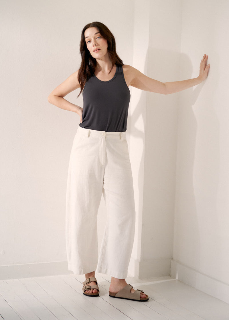 A woman model wearing a grey tank top with white barrel leg jeans, styled with beige sandals, against a beige background