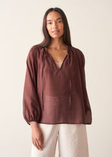 A woman wearing a dark red, brown semi-sheer blouse with a tie necklace and off white trousers.