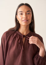 A woman wearing a dark red, brown semi-sheer blouse with a tie necklace and off white trousers.