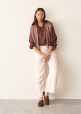 A woman wearing a dark red, brown semi-sheer blouse with a tie necklace and off white trousers.