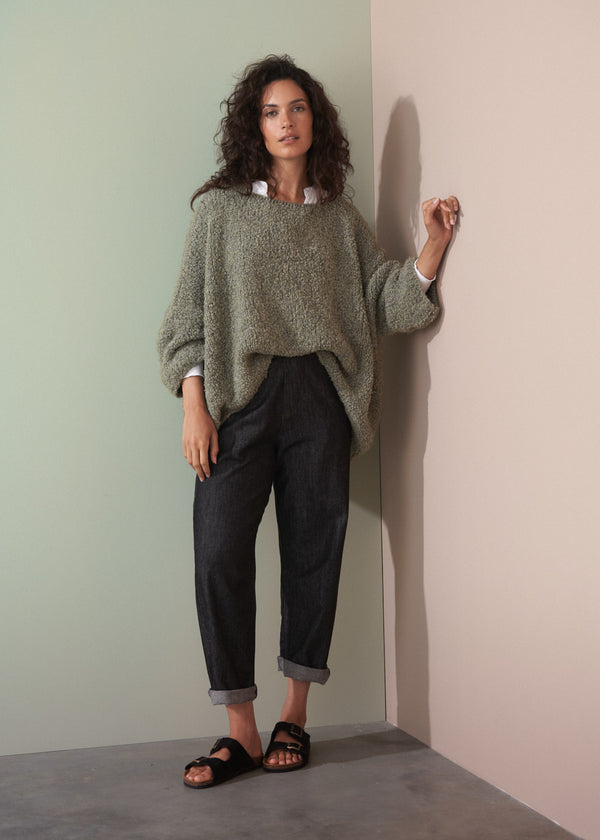 Woman wearing a boucle light green sweater and dark jeans leaning against a plain background
