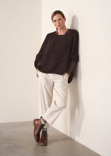 Person wearing a dark boucle sweater and cream trousers against a white wall.