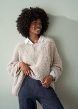 Woman wearing an off white boucle sweater, white shirt and dark blue jeans against a plain background