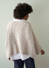 Person wearing an off white boucle sweater, collared shirt and dark blue jeans, facing a plain background