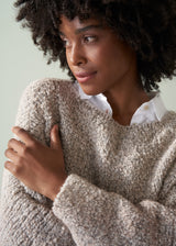 Close up of a woman wearing an off white boucle sweater and dark blue jeans against a plain background