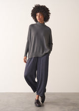 Woman wearing a dark grey turtleneck sweater and blue satin trousers against a plain background