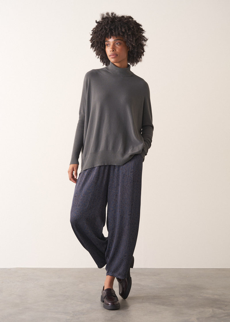 Woman wearing a dark grey turtleneck sweater and blue satin trousers against a plain background