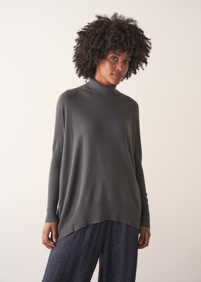 Woman wearing a dark grey turtleneck sweater and blue satin trousers against a plain background