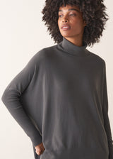 Woman wearing a dark grey turtleneck sweater and blue satin trousers against a plain background