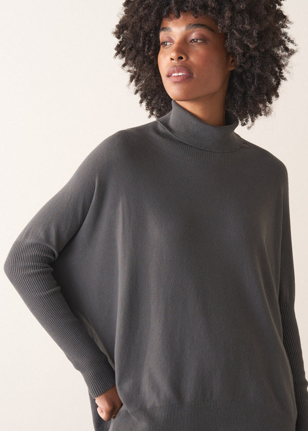 Woman wearing a dark grey turtleneck sweater and blue satin trousers against a plain background
