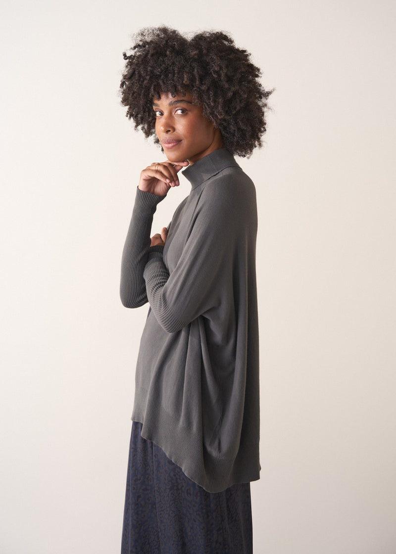 Woman wearing a dark grey turtleneck sweater and blue satin trousers against a plain background