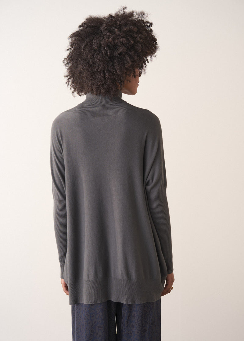 Woman wearing a dark grey turtleneck sweater and blue satin trousers against a plain background