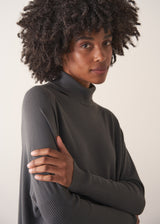 Woman wearing a dark grey turtleneck sweater and blue satin trousers against a plain background