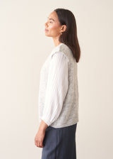 A woman modelling a light grey marled knit waistcoat over a white long-sleeve blouse and navy blue trousers.