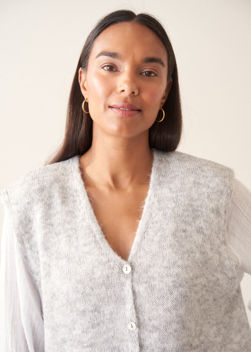 A woman modelling a light grey marled knit waistcoat over a white long-sleeve blouse and navy blue trousers.
