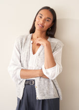 A woman modelling a light grey marled knit waistcoat over a white long-sleeve blouse and navy blue trousers.