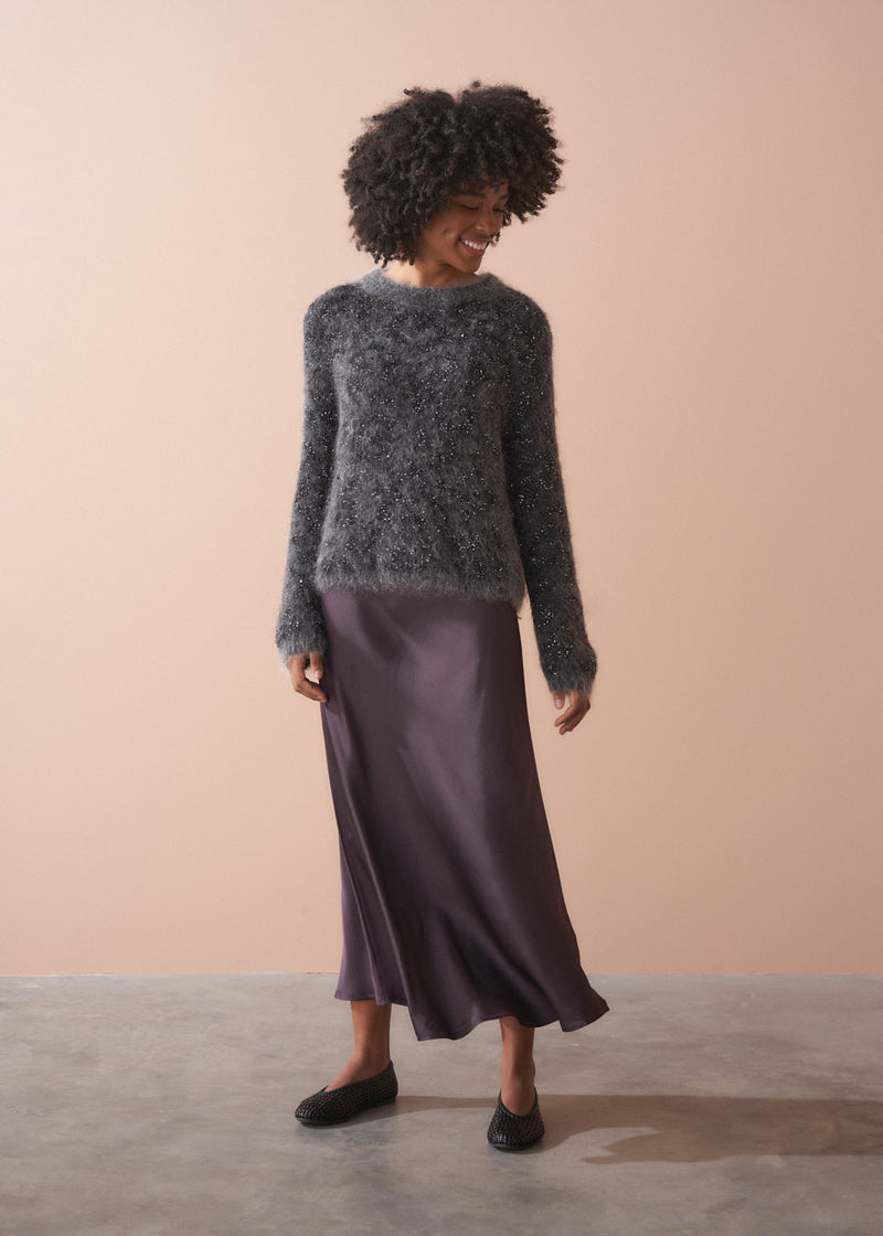 Woman wearing a dark grey fluffy textured sweater with sequin detail, standing against a beige wall