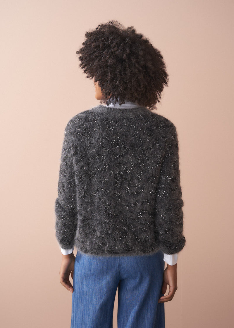 Back of woman wearing a dark grey fluffy textured sweater with sequin detail, blue jeans and collared shirt, facing a beige wall.