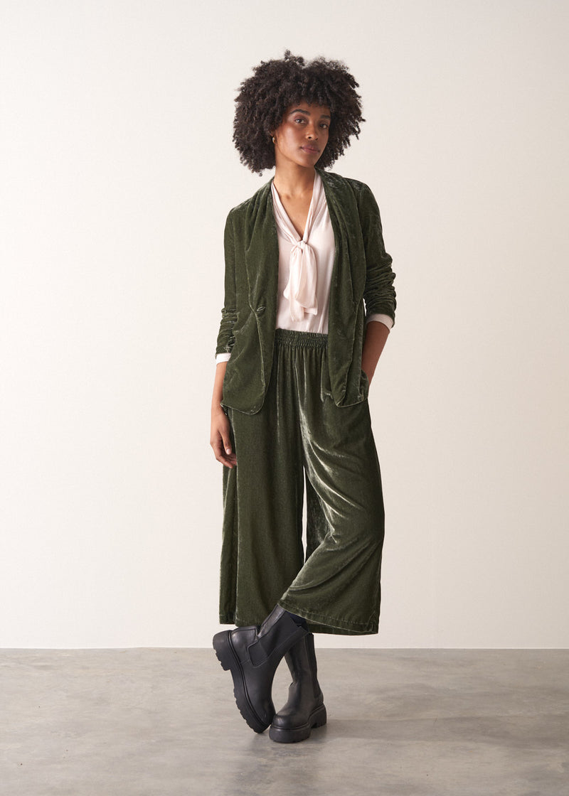 Woman wearing a green velvet blazer and green wide velvet trousers against a white background
