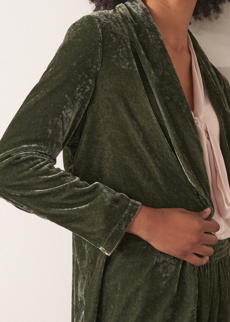 Woman wearing a green velvet blazer and green wide velvet trousers against a white background